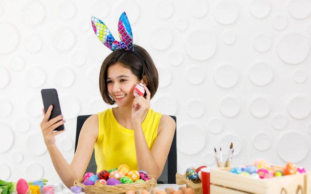 Tavşan kulaklı Asyalı genç kız elinde akıllı telefonla oturmuş gülümsüyor masanın üzerinde renkli Paskalya yumurtalarının resimlerini çekiyor.. - Fotoğraf, Görsel
