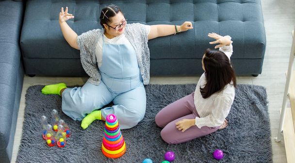 Asian mother sitting on carpet floor in living room smiling playing near a girl with down syndrome daughter in denim overalls with colorful toys. - 写真・画像
