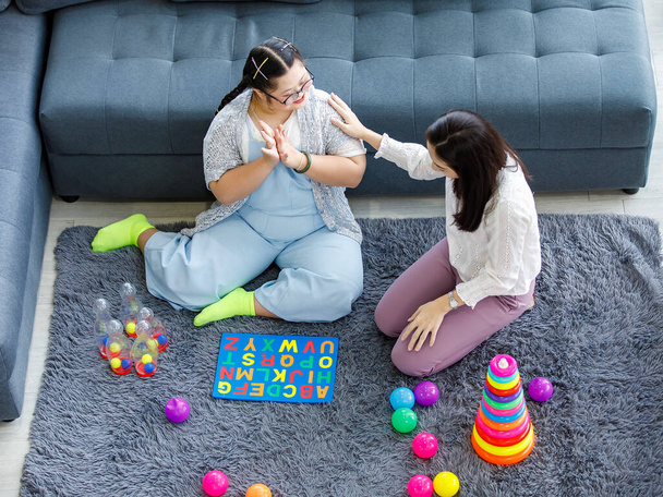 Азиатская мать сидит на полу ковра в гостиной улыбается играя рядом с девушкой с синдромом Дауна дочь в джинсовой комбинезоне с красочными игрушками. - Фото, изображение
