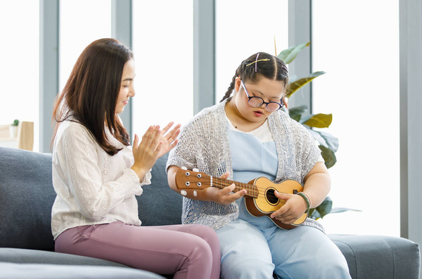 Ασιάτισσα μητέρα κάθεται στον καναπέ στο σαλόνι χαμογελώντας παλαμάκια χέρια διδασκαλία ενθαρρύνει σε ένα κορίτσι με σύνδρομο κάτω κόρη για να μάθουν και να παίξουν μουσική ukulele μικρή κιθάρα στο σπίτι. - Φωτογραφία, εικόνα