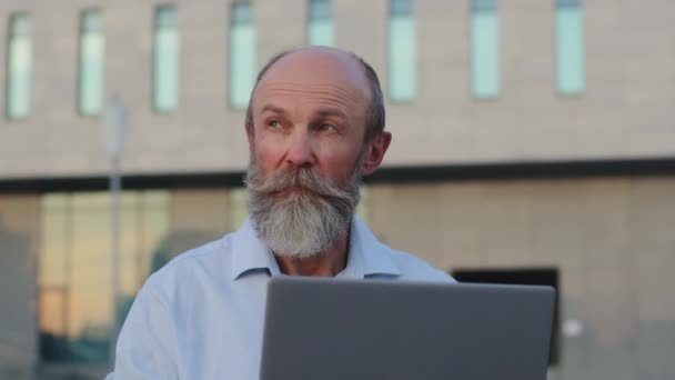 Homme âgé pensif avec barbe grise professionnel créatif travaillant à l'ordinateur portable à l'extérieur regardant de côté, réfléchissant idée. Homme mature pensionné passe du temps libre à l'ordinateur fonctionne à distance envoie des e-mails - Séquence, vidéo