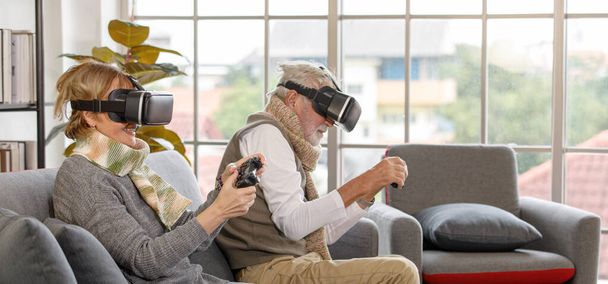 Pareja de ancianos con auriculares VR sentados en el sofá y utilizando gamepads para jugar videojuego contra ventana en la sala de estar en casa. - Foto, imagen