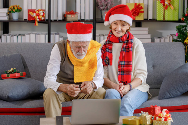 Mature woman and elderly man in warm clothes and Santa hats waving hands while sitting on sofa and making video call on Christmas day in living room at home - Zdjęcie, obraz