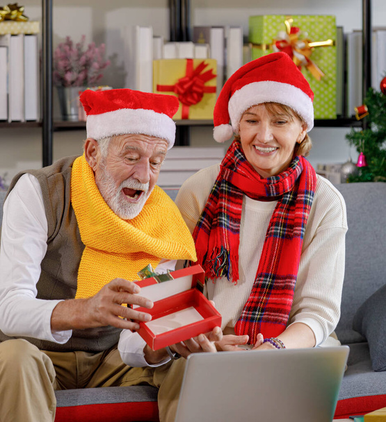 Mujer madura y hombre de edad avanzada en ropa de abrigo y sombreros de Santa saludando las manos mientras se sienta en el sofá y hacer videollamada en el día de Navidad en la sala de estar en casa - Foto, Imagen