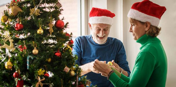 Веселая пожилая пара в шляпах Санта-Клауса улыбается и вешает безделушки на ветки хвойной елки во время подготовки к празднованию Рождества - Фото, изображение