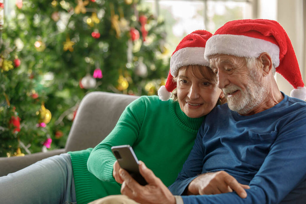 Ευτυχισμένος ηλικιωμένος άνδρας και ώριμη γυναίκα σε Σάντα καπέλα χαμογελώντας και περιήγηση στα μέσα κοινωνικής δικτύωσης στο κινητό μαζί, ενώ κάθεται στον καναπέ κοντά στο χριστουγεννιάτικο δέντρο - Φωτογραφία, εικόνα