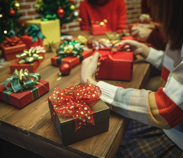 Krásná dárková krabička zabalená a zabalená rodinnou skupinou žen na svetru s barevným papírem, stuhou, půvabnou mašlí zdobit luxusní vánoční stromek pro šťastnou přípravu zimní párty v prosinci - Fotografie, Obrázek