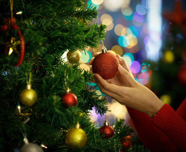 Χέρι της γυναίκας σε santa πουλόβερ κρέμεται πολύχρωμο γοητευτικό στολίδι και μπάλα στο χριστουγεννιάτικο δέντρο για να διακοσμήσετε πανέμορφο πάρτι γιορτή την ευτυχισμένη νύχτα. Προσθέστε κάποιο θόρυβο για να χωρέσει vintage-style εικόνα. - Φωτογραφία, εικόνα