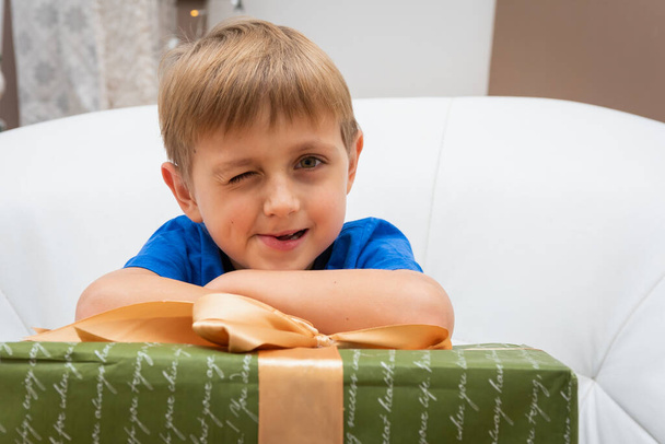 Ευτυχισμένο ξανθό αγοράκι έσκυψε τα χέρια του σε ένα χριστουγεννιάτικο δώρο, κάθεται σε μια ελαφριά δερμάτινη καρέκλα κοντά στο χριστουγεννιάτικο δέντρο με ένα πακεταρισμένο κουτί δώρου στην αγκαλιά του και χαμογελώντας στην κάμερα - Φωτογραφία, εικόνα