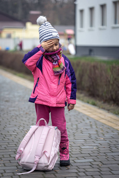 Маленька дівчинка з рюкзаком, в куртці і капелюсі біля школи
. - Фото, зображення