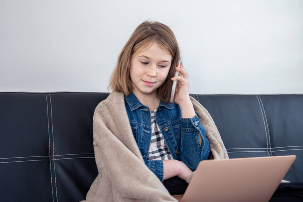 Uma adolescente está sentada no sofá em um laptop e falando ao telefone. - Foto, Imagem