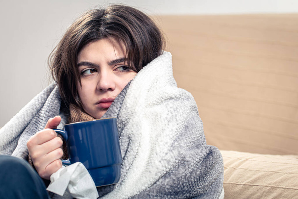 病気の若い女性が毛布に包まれたお茶を飲みながら座っています. - 写真・画像