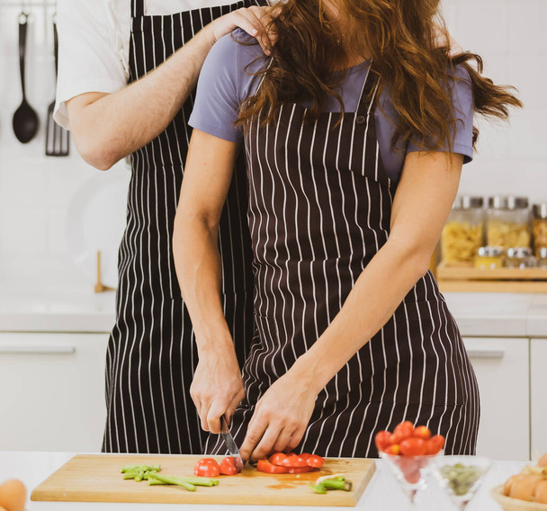 Sevgilisinin yanında duran olumlu kız arkadaş evdeki hafif mutfakta çeşitli ürünlerle masada elma kesiyor. - Fotoğraf, Görsel