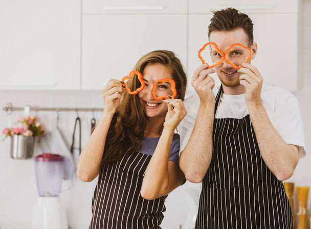 Весела пара в фартухах дивиться на камеру через скибочки червоного болгарського перцю, стоячи на світлій сучасній кухні
 - Фото, зображення