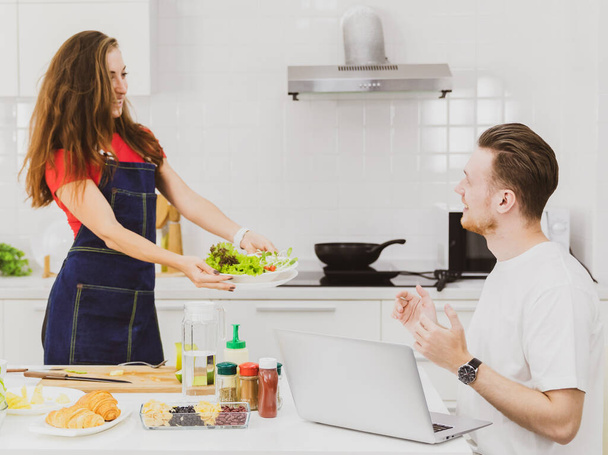 Vista lateral de la novia en delantal mostrando tazón de ensalada fresca para novio sorprendido cerca de la mesa con productos en la cocina - Foto, imagen