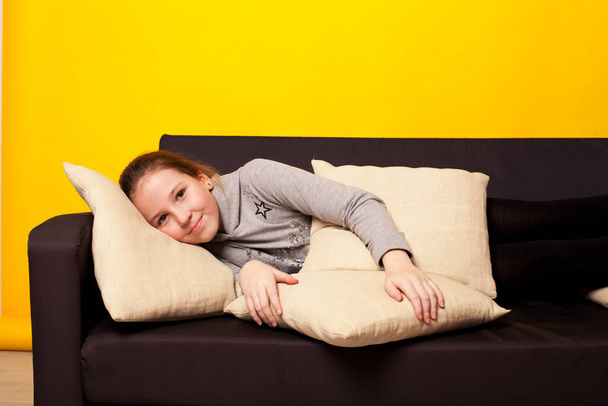 niña en un sofá negro en casa sobre un fondo amarillo - Foto, Imagen