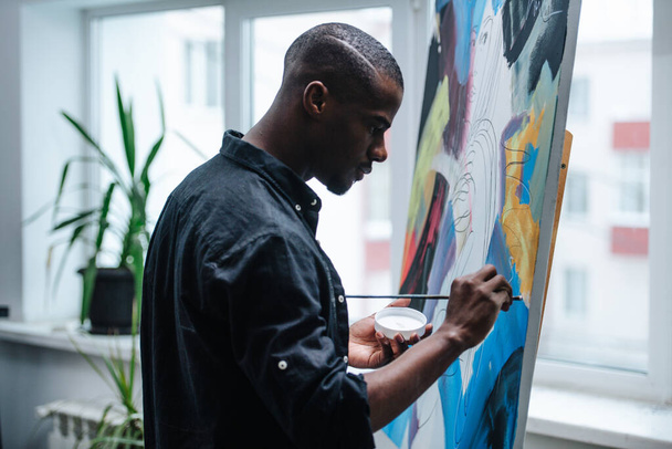 Side view of a black man painting on an easel inside of his apartment. He is coloring it. Window view on the building next door. - Foto, Imagen