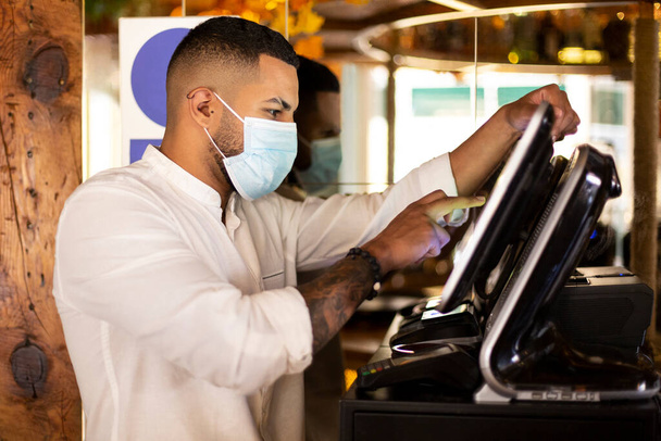 Νεαρός Λατινοαμερικάνος υπάλληλος εστιατορίου με ιατρική μάσκα προετοιμάζει το λογαριασμό στο ταμείο του μηχανήματος.  - Φωτογραφία, εικόνα