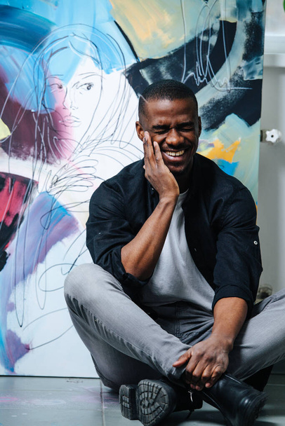 Laughing african american sitting in front of an abstract painting of a woman. Sitting cross-legged, leaning on his hand, eyes closed. - Foto, Imagen