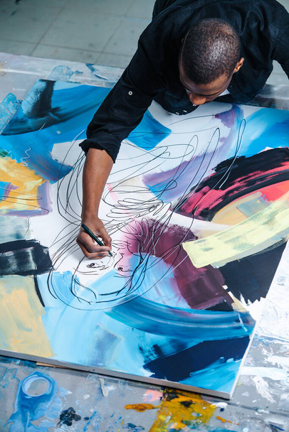 High angle image of a black man drawing lines on a big canvas with a painting on a table, leaning over it. He is holding a felt-tip pen. Inside of an apartment. - Photo, image