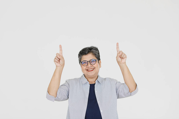 Оптимистичная азиатка средних лет в очках, смотрящая на камеру с улыбкой и указывая на серый фон - Фото, изображение
