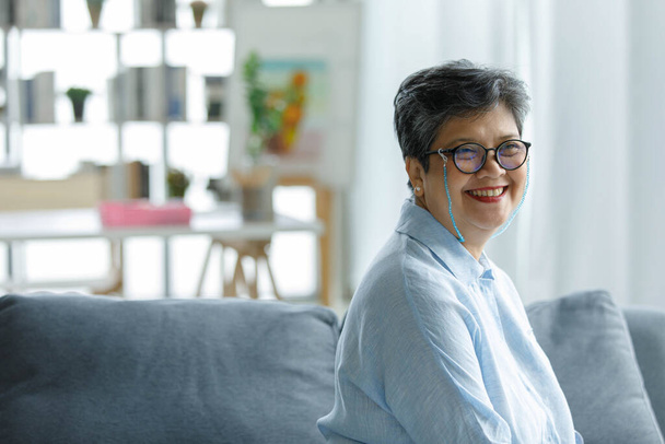 Side view of happy mature Asian female in glasses smiling and looking at camera while sitting on couch at home - Foto, afbeelding