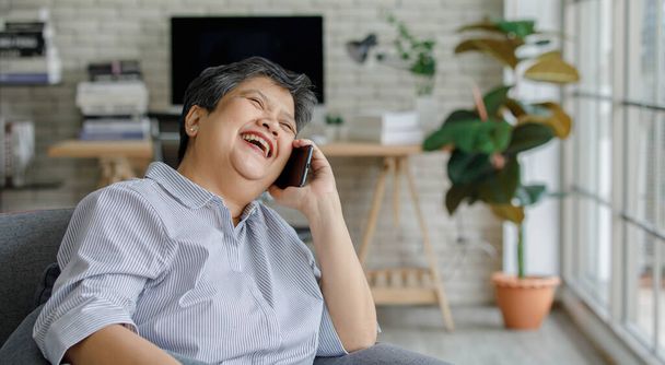 Pozytywny w średnim wieku Azjatka uśmiecha się i odwraca wzrok siedząc na kanapie i rozmawiając na smartfonie we współczesnym salonie w domu - Zdjęcie, obraz