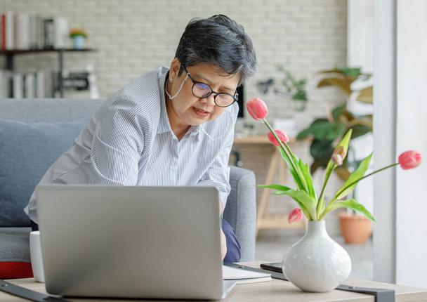 Азиатка средних лет в очках пишет в планировщике рядом с ноутбуком и вазой с тюльпанами, сидя на диване и работая над удаленным проектом дома - Фото, изображение