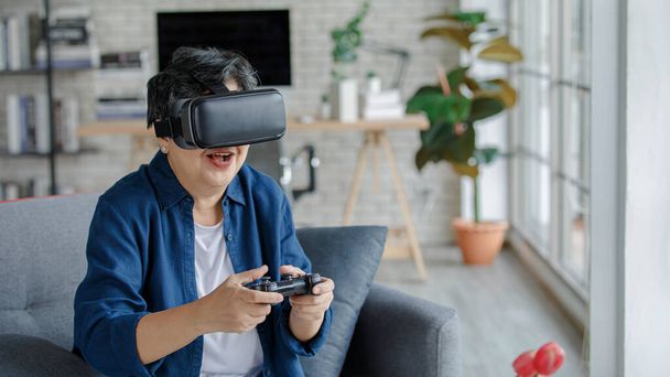 Middle aged female in VR headset smiling and playing videogame while sitting on sofa on weekend day in living room at home - Fotó, kép