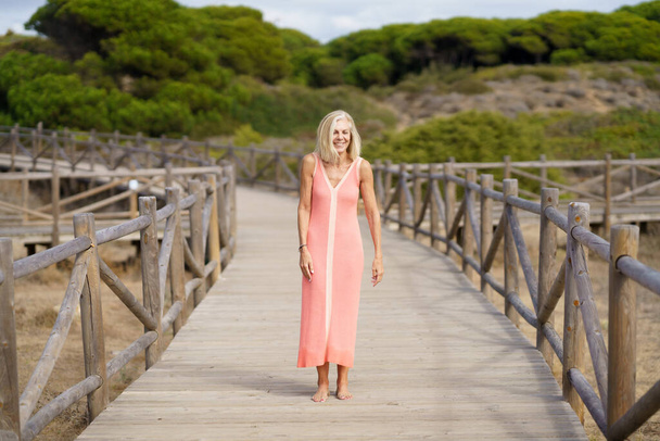 Hermosa mujer madura caminando a lo largo de un camino de madera cerca de la playa., vistiendo un bonito vestido naranja. - Foto, imagen