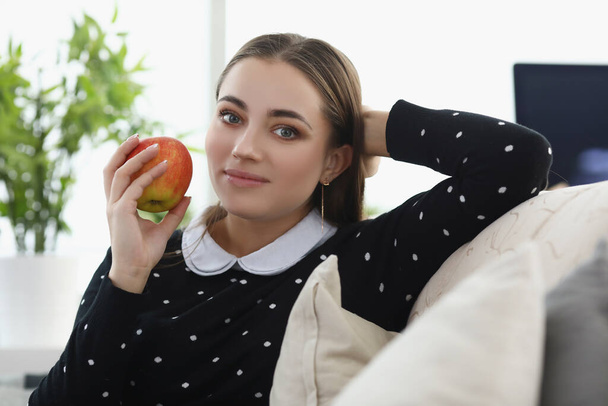 Bonita joven sentada cómoda en el sofá en casa, fruta fresca de manzana como merienda - Foto, imagen