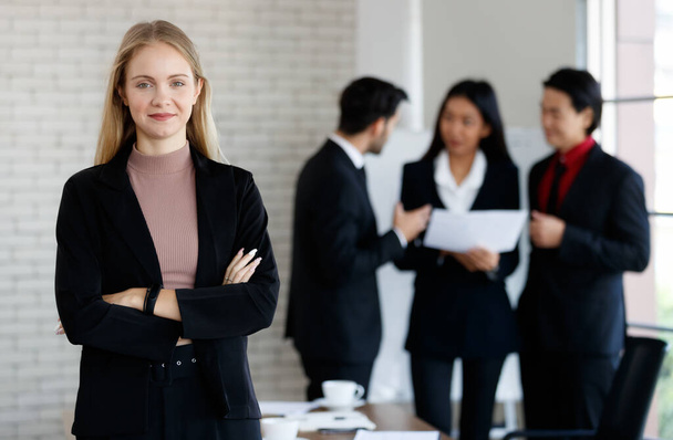 Positive female entrepreneur in elegant outfit looking at camera while standing near group of multiracial coworkers in light modern conference room - Photo, image