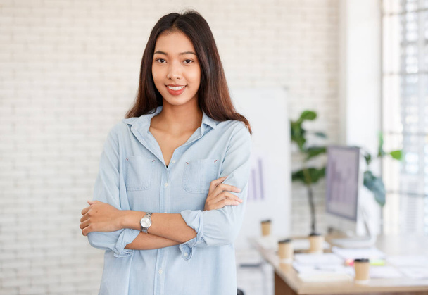 Positieve Aziatische vrouwelijke manager met lang haar en gekruiste armen met elegante outfit kijkend naar de camera terwijl hij in een lichte moderne werkruimte staat - Foto, afbeelding
