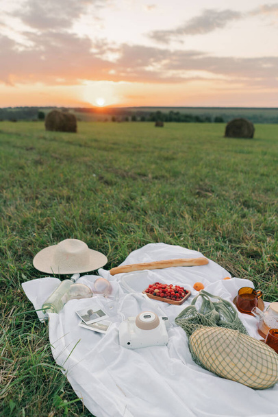 Tablecloth with fruits, picnic food, hat and film camera in the middle of a field at sunset - Photo, Image