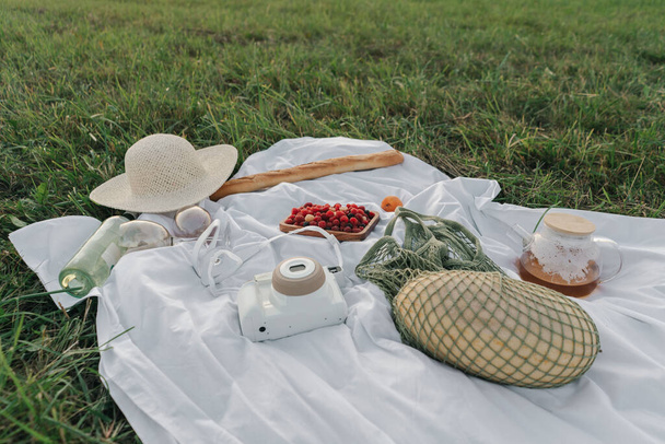 Gün batımında tarlanın ortasında içinde meyve, piknik yemeği, şapka ve film kamerası olan masa örtüsü. - Fotoğraf, Görsel