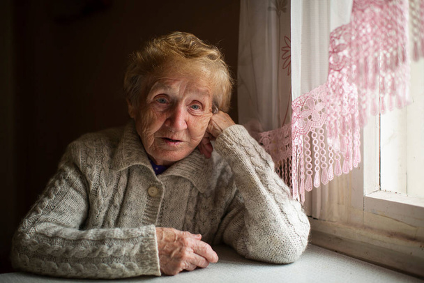 Järkyttynyt ja hämmentynyt vanha nainen istuu ikkunassa talossaan.. - Valokuva, kuva