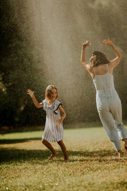 Cute little girl having fun with water under irrigation sprinkler with her mother - Foto, Imagem