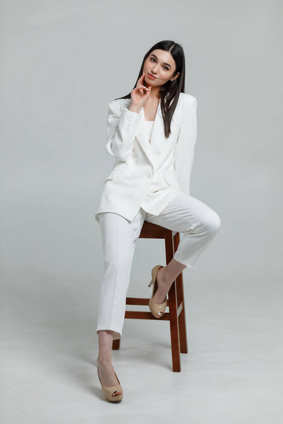Foto einer schönen brünetten Modell trägt weißen Business-Anzug lächelnd und Blick in die Kamera an einem Stuhl - Foto, Bild