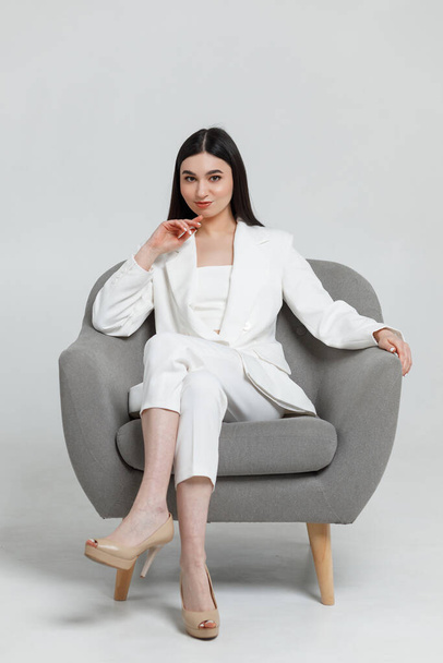 Foto de uma bela cosmetologista morena vestindo terno branco sentado em uma cadeira cinza e sorrindo com a mão perto de sua cabeça - Foto, Imagem