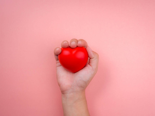 Hand holding and squeezing red heart ball on pink pastel background. Concept of love, care, giving, charity, health insurance, well-being, minimal style. - Фото, изображение