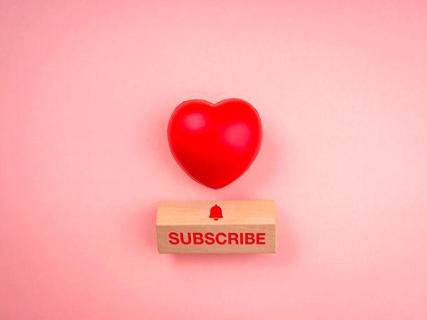 Abonneer concept. Word "SUBSCRIBE" en bel pictogram op houten kubusblok en rode hart bal op roze pastel achtergrond, minimale stijl. Sociale media volgen. - Foto, afbeelding