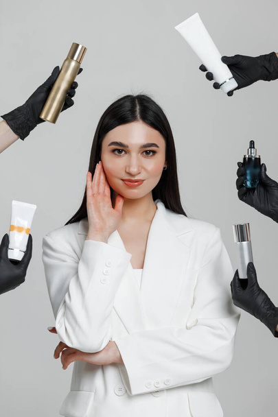 Foto einer gut aussehenden Kosmetikerin, die in einem Studio posiert, mit Händen in schwarzen Handschuhen, die Kosmetikprodukte um sich halten - Foto, Bild