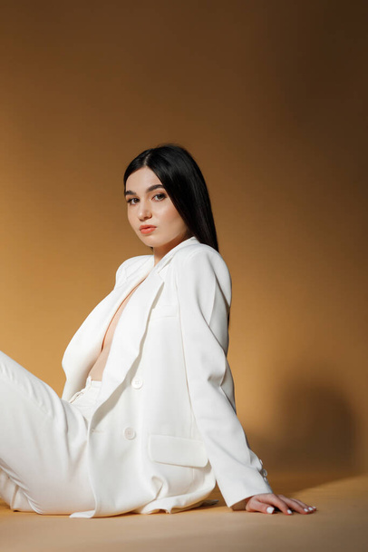 Photo of a young pretty woman wearing a white suit while posing in a studio with orange background looking at camera - Foto, immagini