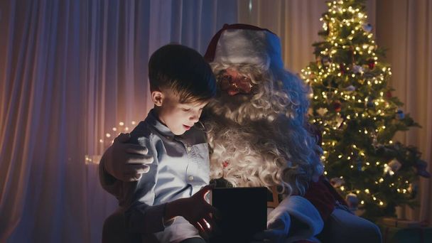 Little cute boy sitting on Santas knees, opening a gift in front of Christmas tree. Adorable child is happy and surprised. - Photo, image
