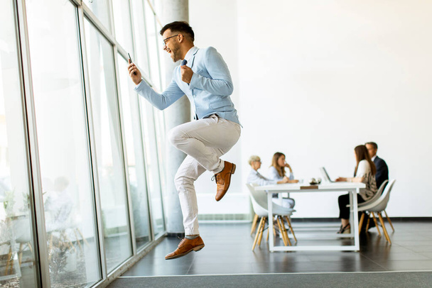 Handsome young businessman with smartphone in a modern office jumping after receving a great news - Foto, afbeelding