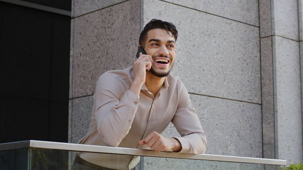 Portrait of cheerful smiling carefree happy hispanic arabic bearded business man boss leader guy standing on balcony terrace talking on mobile phone answering call talking wireless smartphone laughing - 写真・画像