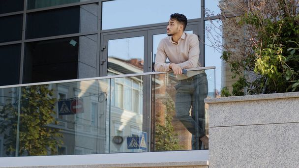Confident young hispanic business man standing at railing modern skyscraper office building looking away holding phone thinking resting. Successful leader guy male on balcony terrace relaxing think - Valokuva, kuva