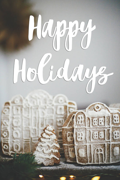 Happy Holidays text on christmas gingerbread cookie houses and trees with golden lights on rustic table. Season's greetings card. Handwritten sign. Merry Christmas! - Foto, Bild