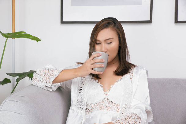 Mujer asiática en camisón blanco bebiendo agua tibia en la cama en la sala de estar. - Foto, imagen