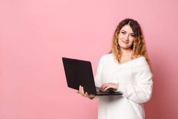 A cheerful young woman standing isolated on a pink background using a laptop computer and looking at the camera. - Foto, Bild